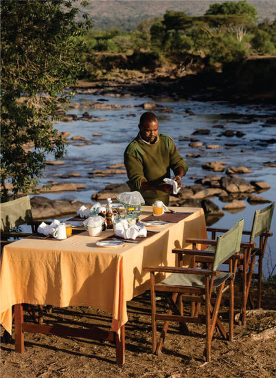 Man setting up a table for bush dinner near River to enjoy a unique dining experience on group accommodation in Masai Mara at Budget Lodge