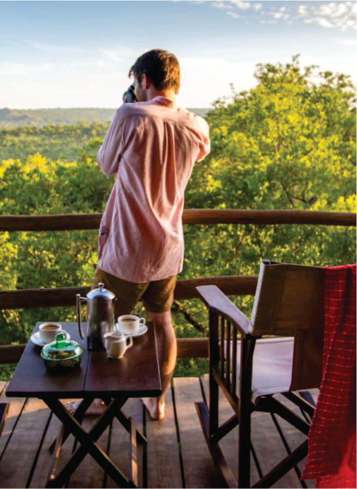 Man standing and taking a photo on terrace near chair and breakfast table at Masai Mara budget camp