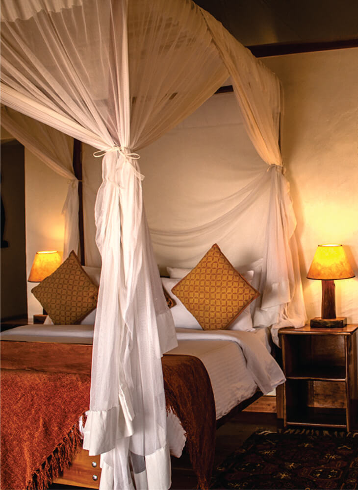 Short-stay accommodation Bedroom at Lion Sands Masai Mara Lodge with a wooden bed, mosquito net, and two bedroom lamps in the corner next to it